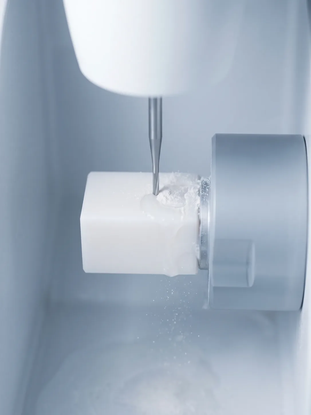 Interior de una fresadora: una fresa talla un bloque cerámico blanco y levanta polvo
